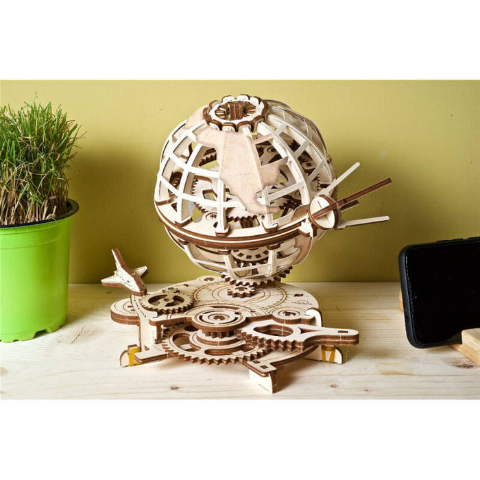 Intricate light wood mechanical 3D globe puzzle with gears and orbiting satellites next to a small green plant.