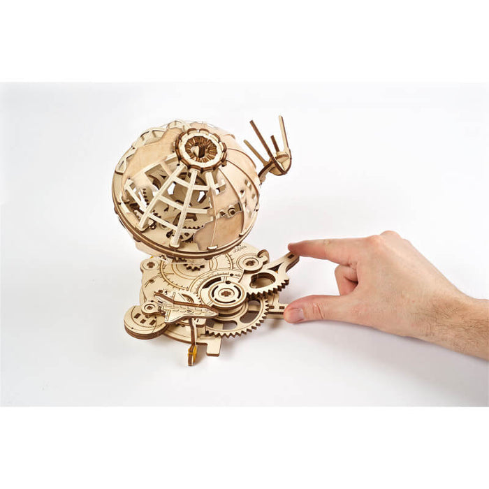 Hand operating an intricate wooden mechanical globe puzzle with gears, a satellite, and a space shuttle.