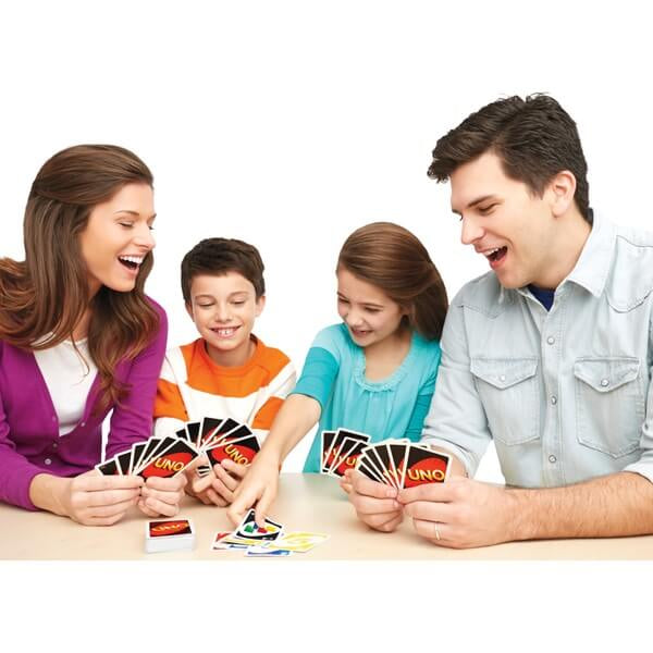 A smiling family of four plays the Uno card game with colorful cards around a light-colored table.