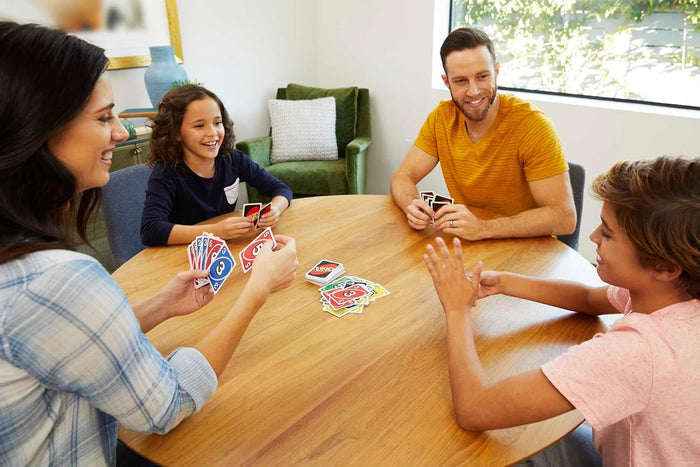 Family playing Uno card game with colorful red, blue, yellow, and green cards on a round wooden table.