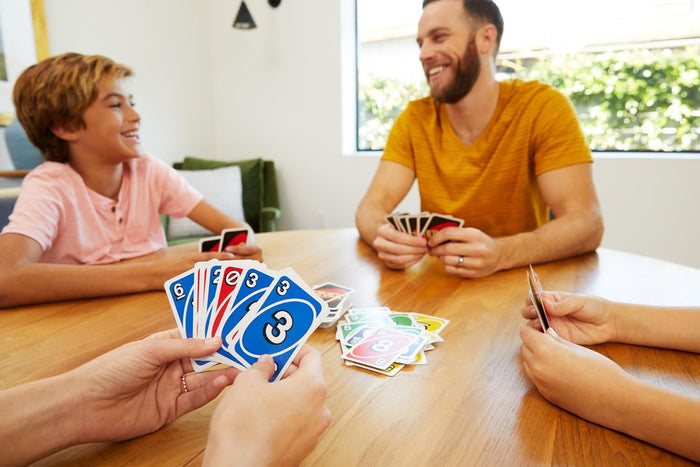 Hands hold blue and red numbered cards over a pile of colorful Uno cards during a family game at a wooden table.