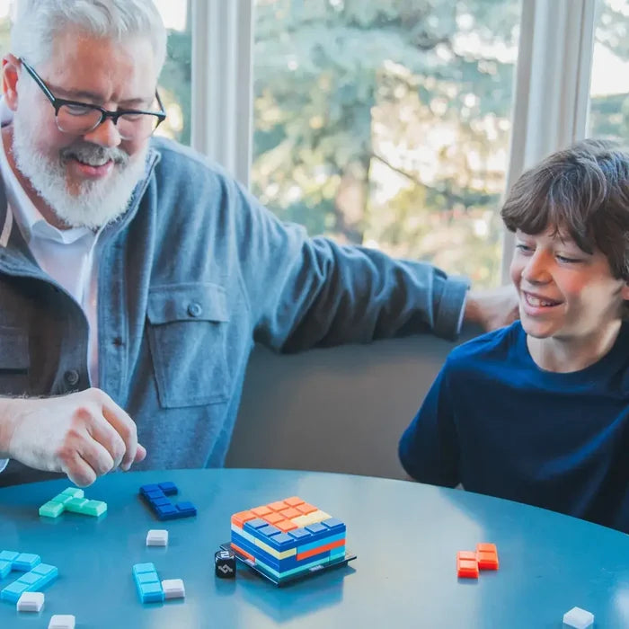 Man and boy play a game stacking colorful, interlocking 3D blocks to form a cube on a blue table.