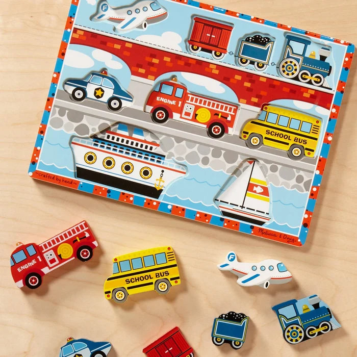Colorful wooden puzzle board with chunky bus, fire truck, and plane pieces scattered on a light wood surface.