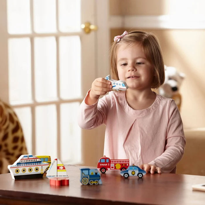 Young girl playing with chunky wooden vehicle puzzle pieces like an airplane, fire truck, and boat on a table.
