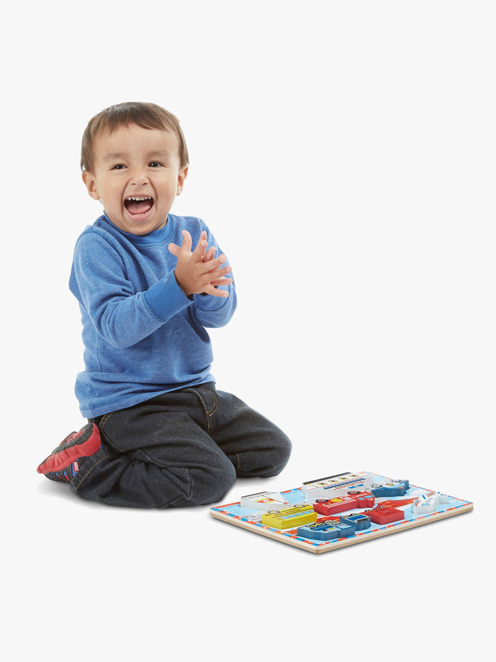 A young boy laughs and claps next to a colorful wooden puzzle with chunky vehicle-shaped pieces.