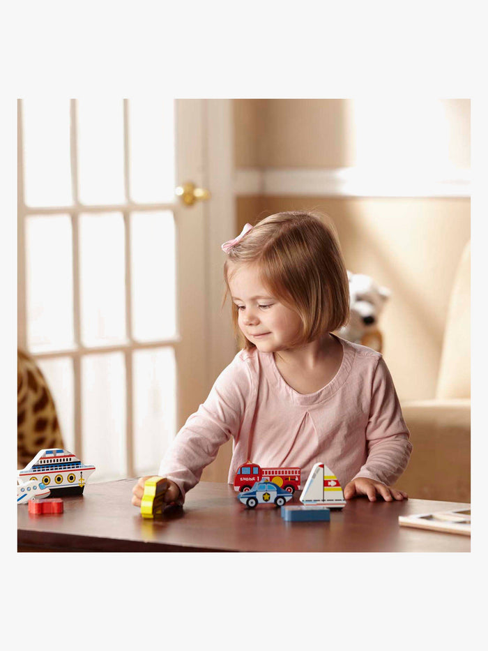 A toddler plays with chunky wooden vehicle puzzle pieces like a red fire truck, blue police car, and white cruise ship.