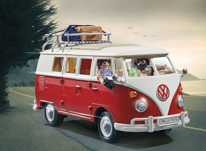 Red and white toy camper van with two figures and luggage on a silver roof rack, shown on a coastal road.