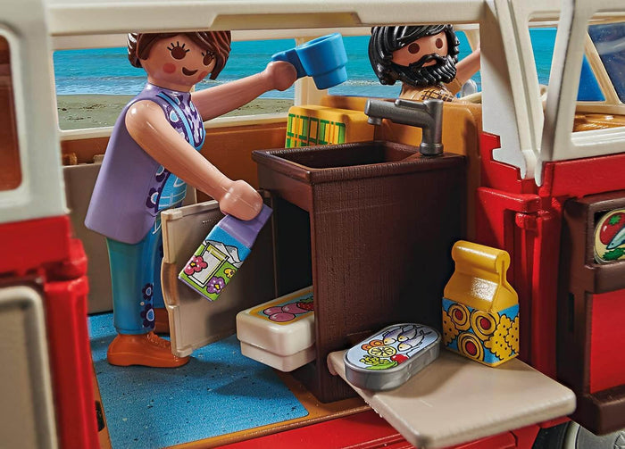 Interior of a red toy camper van with two figures, a sink, and tiny food accessories against a beach background.