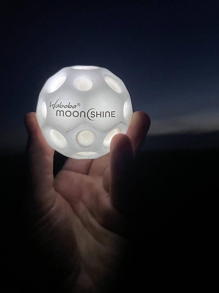 A hand holds a glowing white Waboba Moonshine ball with crater indentations against a dark night sky.