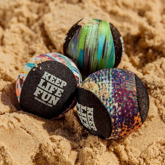 Three colorful, fabric-covered water balls with Keep Life Fun text resting on a sandy beach.