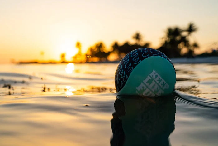 Teal and navy patterned ball with KEEP LIFE FUN text floating in calm water during a golden sunset.