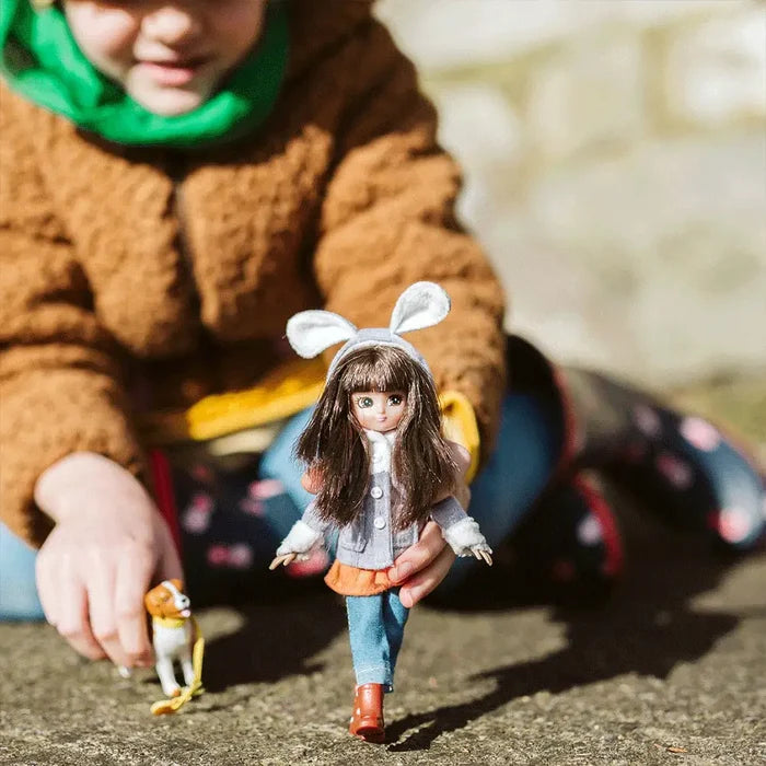 Doll with brown hair, bunny ears, and a grey jacket held by a child outdoors next to a small toy dog.