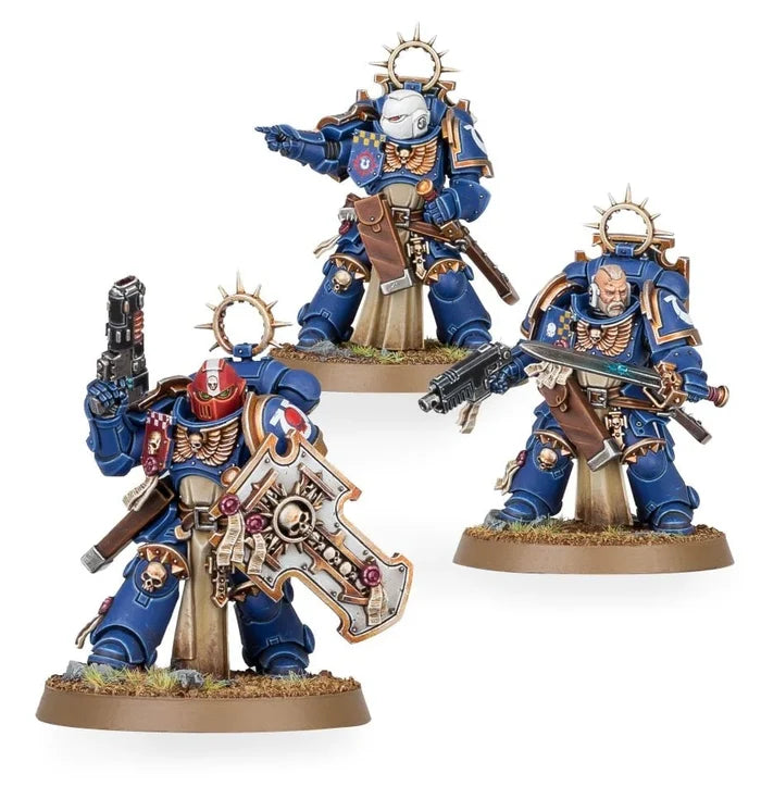 Three blue Space Marine miniatures in ornate gold trim armor with power swords, a storm shield, and tan tabards.