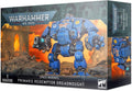 Blue armored robot miniature with heavy weaponry on a box featuring fiery ruins and a dark sky with faint faces.