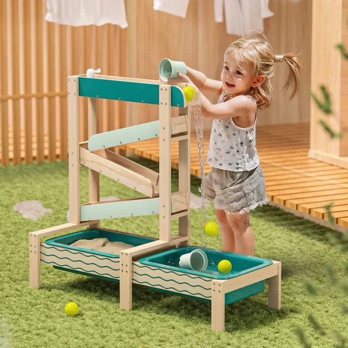 Child pouring water into a tiered wooden water track toy with teal basins and yellow balls on a grassy lawn.