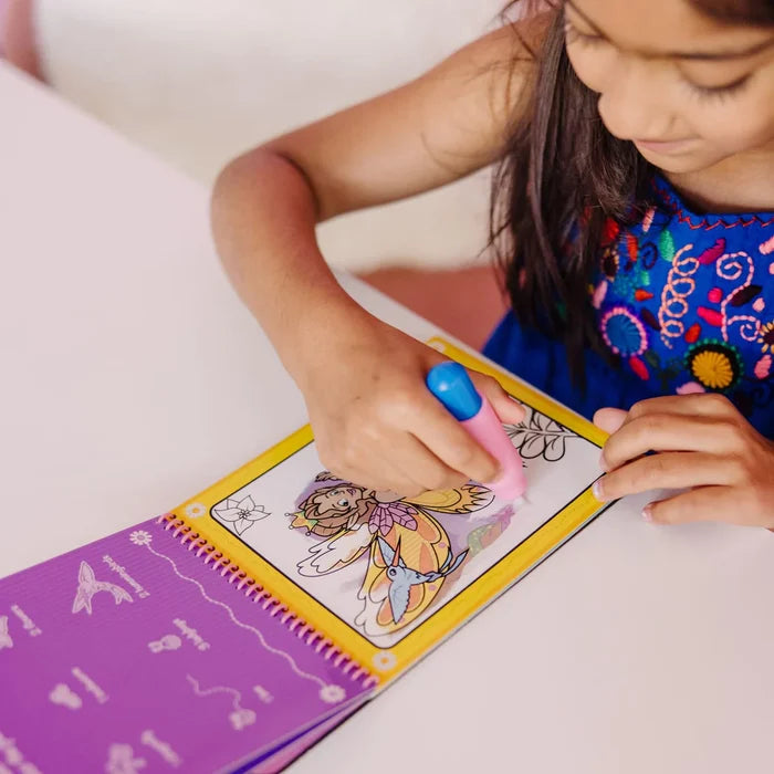 Child using a pink and blue water pen to color a fairy scene in a yellow spiral-bound travel activity book.