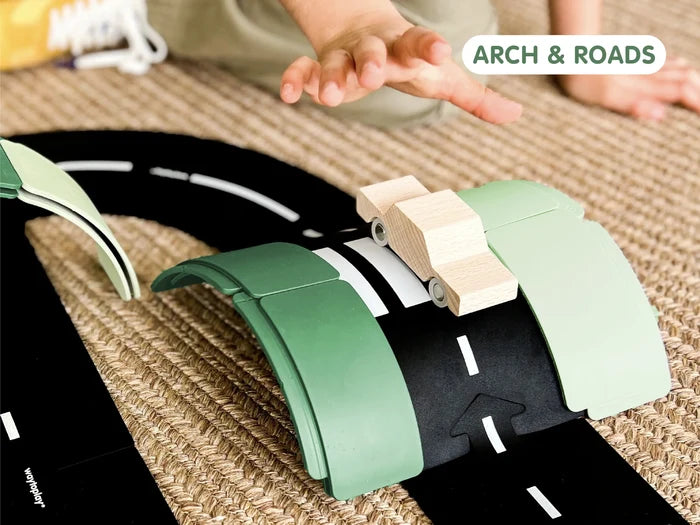 Child reaching for a wooden car on a black flexible road arched over green blocks on a tan woven rug.