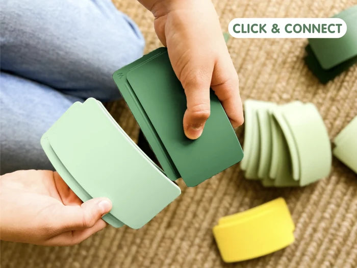 Hands connecting light and dark green curved building pieces from a toy set on a textured rug.