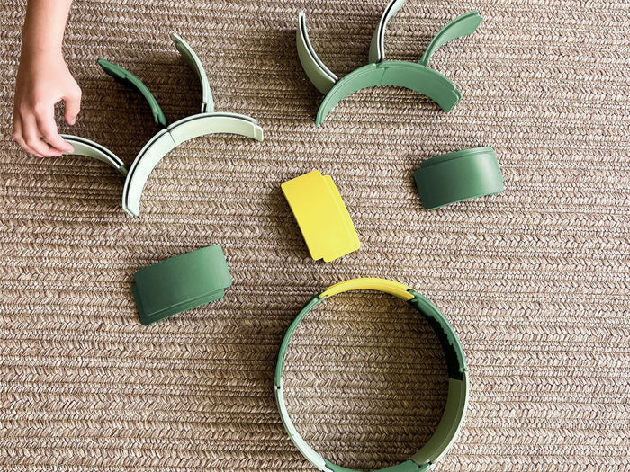 Child's hand arranging green and yellow arched plastic building pieces on a brown textured woven rug.