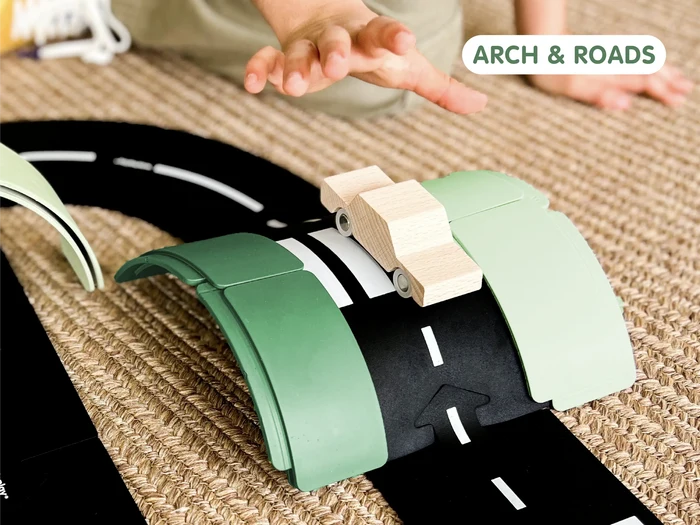 Wooden toy car on a black flexible road track arched over green plastic pieces on a textured rug.