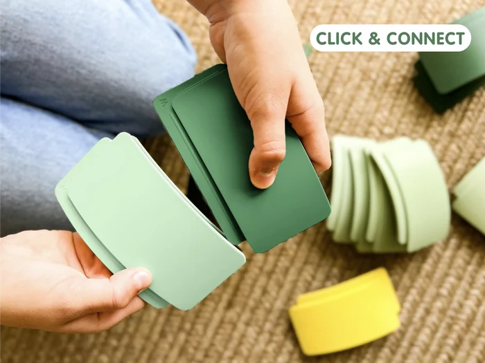 Child's hands click together light and dark green arched toy pieces over a textured rug with colorful curved blocks.