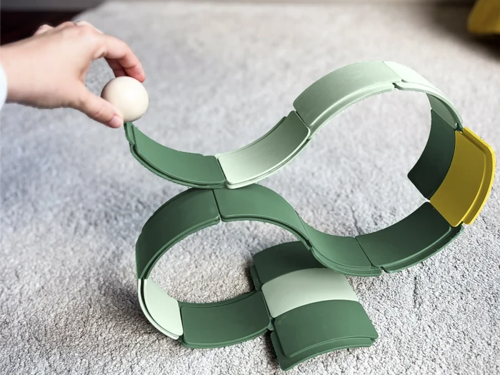 A hand places a wooden ball on a wavy track made of interlocking green and yellow arched pieces on a gray carpet.