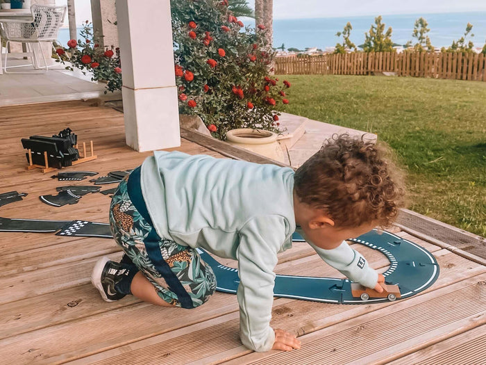 Toddler playing with black flexible racing road tracks and a wooden toy car on an outdoor wooden deck.