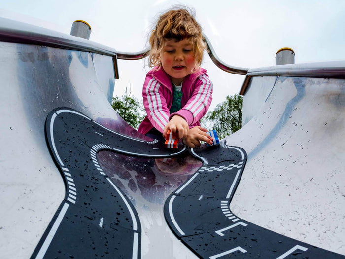 A child plays with a flexible black toy race track and small toy cars on a wet metal playground slide.
