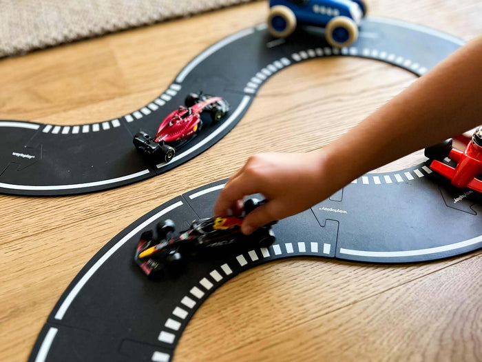 Child plays with toy race cars on interlocking black rubber tracks with white racing lines on a wooden floor.