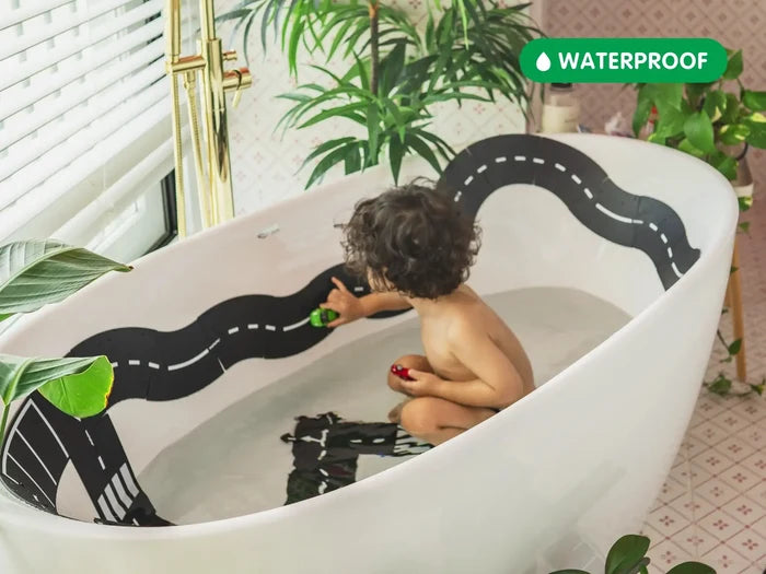 Child in a bathtub playing with black flexible road tracks with white dashed lines stuck to the side of the tub.