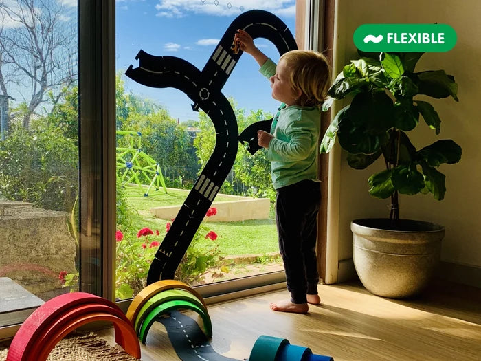 A toddler plays with a flexible black rubber toy road track stuck to a glass door. Wooden rainbow arches sit on the floor.