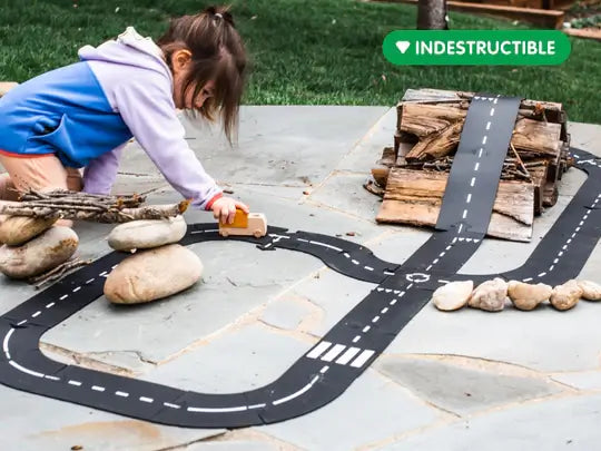 A child plays with a flexible black toy road set and wooden car on an outdoor stone patio with logs and rocks.