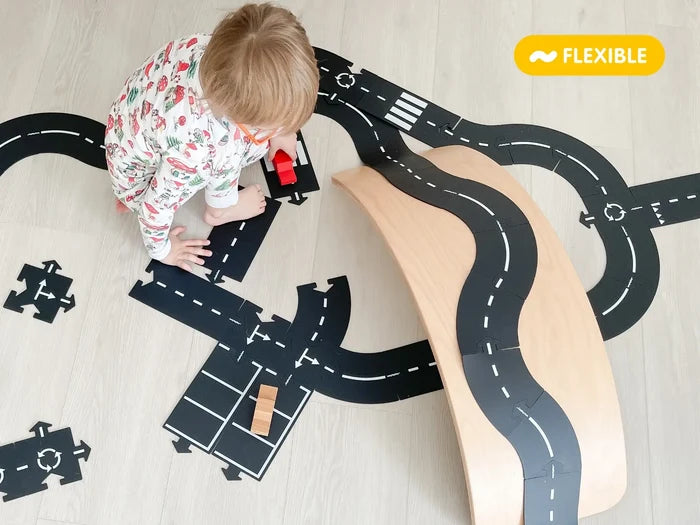 Toddler playing with flexible black rubber road tracks that curve over a wooden ramp on a light floor.
