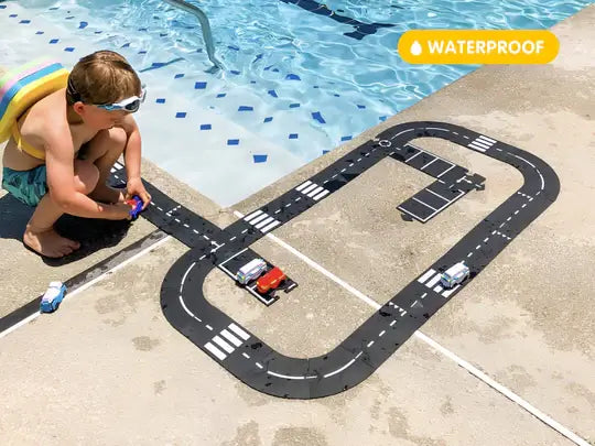Child plays with modular black road pieces and cars on a pool deck. A yellow waterproof sticker is in the corner.
