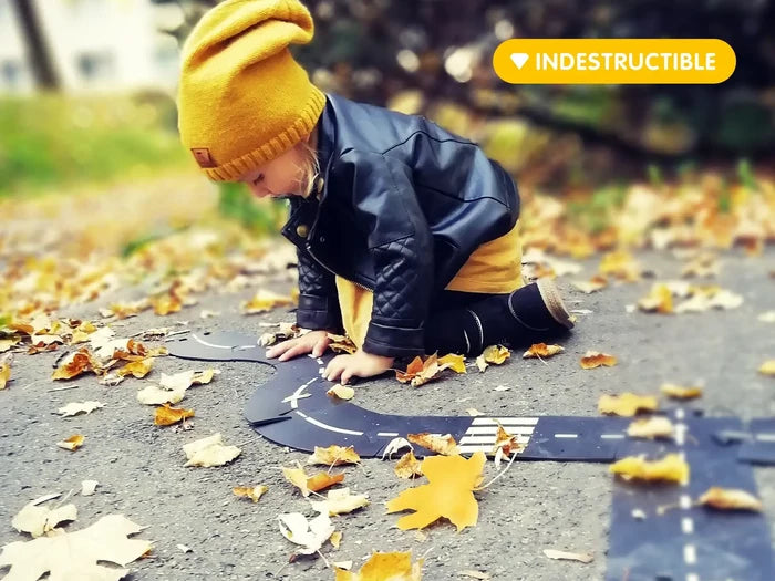 Child plays with flexible black toy road tracks on leafy path. Image includes yellow INDESTRUCTIBLE banner.