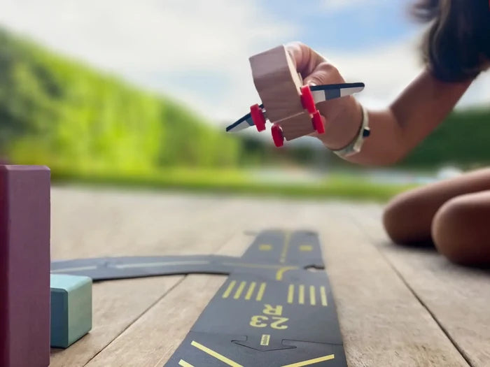 Hand holds a wooden toy plane over flexible black runway tracks with yellow airport markings on a wooden deck.