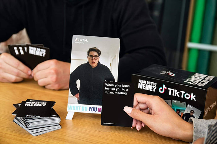 People playing a card game with TikTok photo and text cards on a wooden table next to the black and white game box.