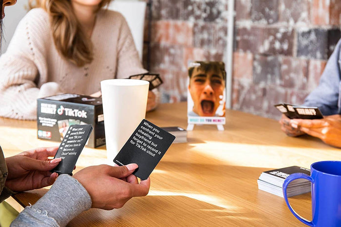 Hands hold black caption cards next to the TikTok game box and a screaming photo card on a sunlit wooden table.