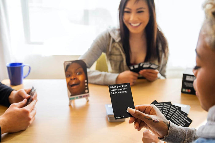 Friends playing a card game with black caption cards and a shocked meme photo card on a small display easel.