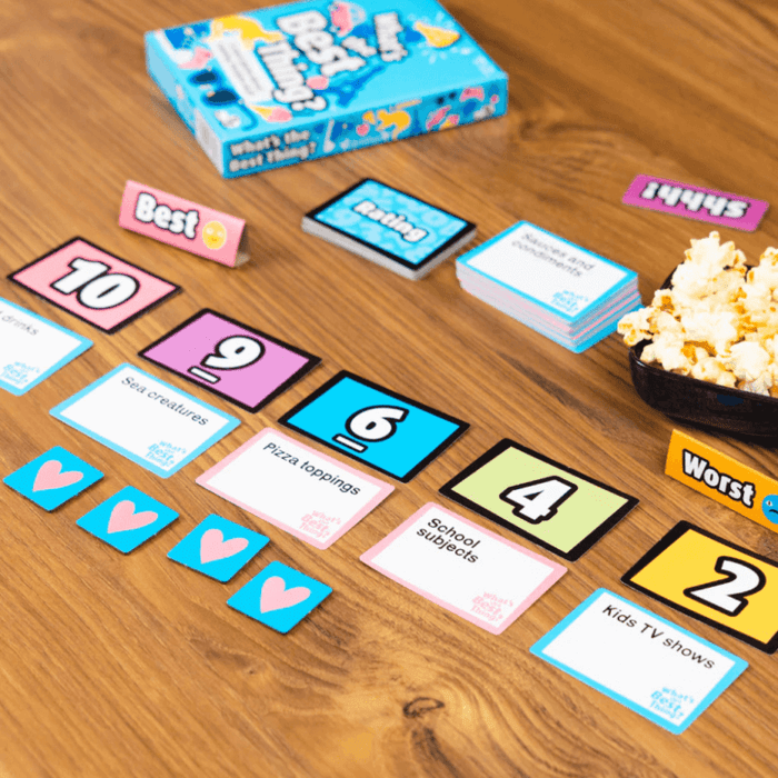 Blue board game box with colorful numbered cards, category tiles, and heart tokens on a wood table with popcorn.