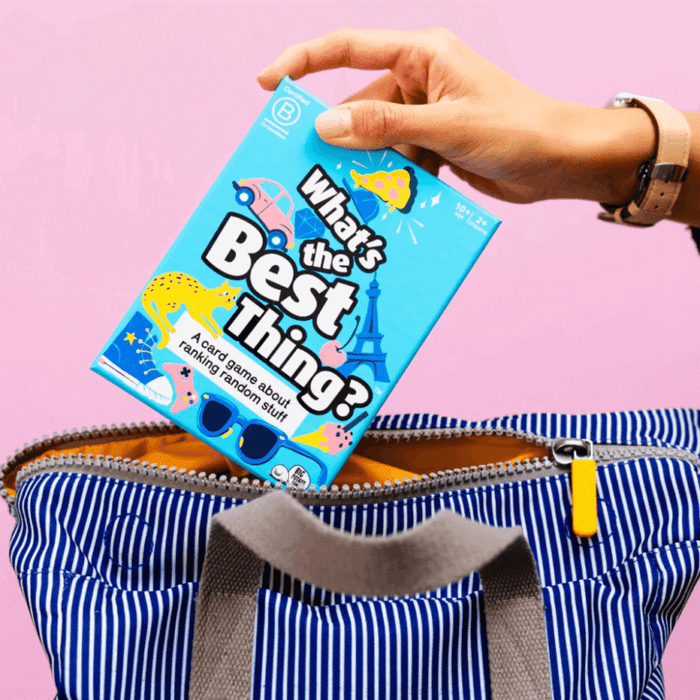 Hand placing a bright blue What's the Best Thing? game box with colorful icons into a striped bag on a pink background.