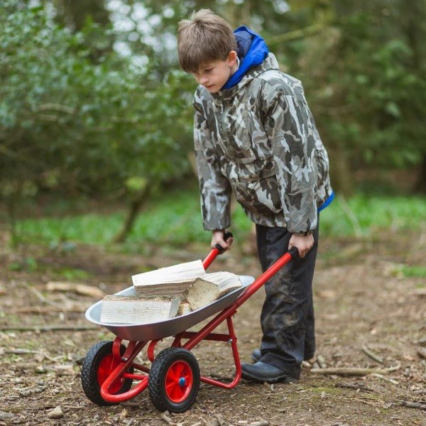 Child pushing a red metal two-wheel toy wheelbarrow with a grey tray filled with wood logs in a forest.