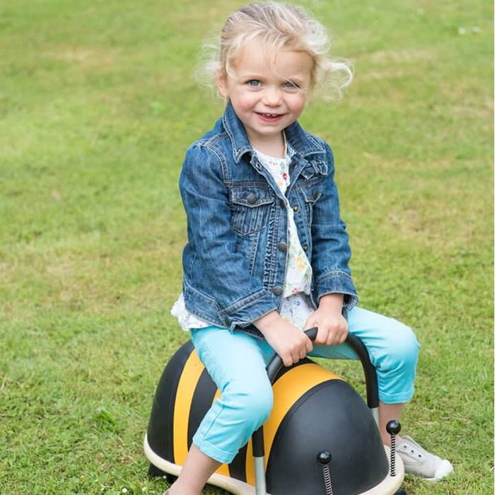 Child riding a yellow and black striped bee toy with black handles and springy antennae on a grassy lawn.