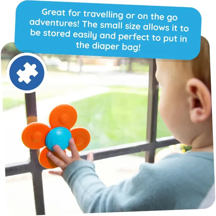 Toddler spins an orange four-petal flower suction toy with a blue center on a glass window.