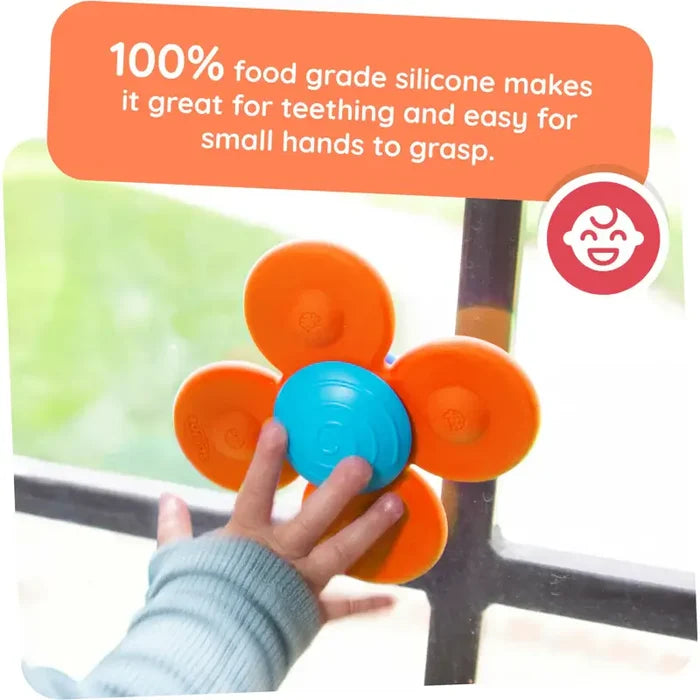 A baby's hand reaching for a flower-shaped orange and blue silicone suction toy attached to a window.