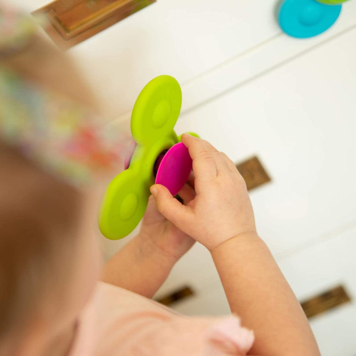 Toddler hands holding a lime green three-lobed spinning toy with a pink suction cup base against a white surface.