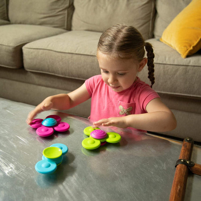 A child plays with three colorful, flower-shaped silicone suction cup fidget spinners on a shiny metal table.