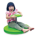 A child sits and laughs on a bright green spinning toy with a large circular base and a small top handle disk.