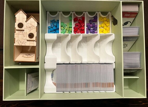 Light green game box with a wooden birdhouse tower, colorful tokens in white trays, and sorted stacks of cards.