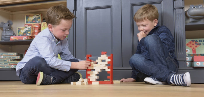 Two boys play with a red wooden frame game, removing tan blocks from a stacked wall on a light-colored floor.